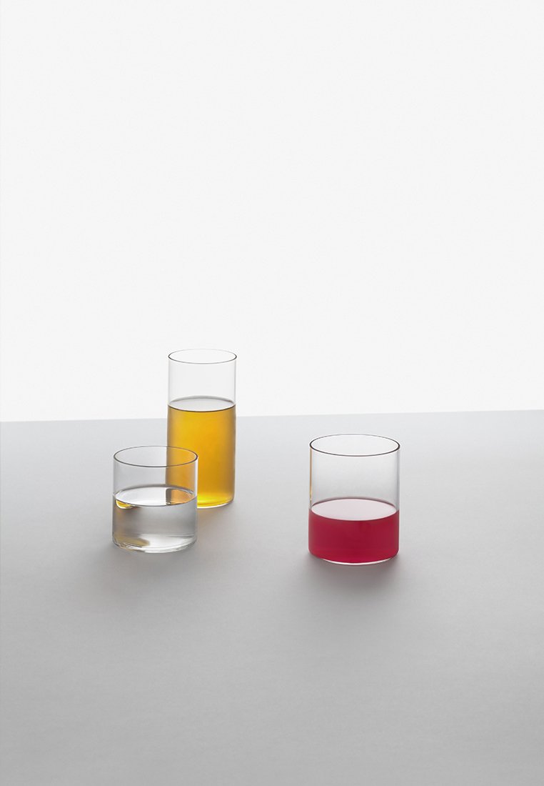 Trzy przejrzyste szklanki na szarej powierzchni, każda zawiera inny płyn: wodę, bursztynowy płyn i czerwony płyn, różniące się wysokością i poziomem napełnienia.