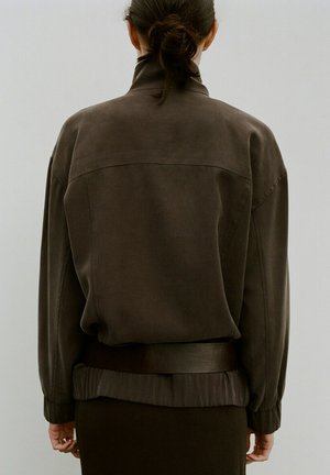 Femme aux cheveux foncés attachés en chignon, portant une veste oversize marron foncé avec une large ceinture, debout de dos sur un fond uni.