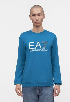 Homme aux cheveux courts portant une chemise bleue à manches longues avec le logo blanc « EA7 Emporio Armani » et un jean bleu sur un fond uni.