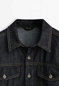 Ciemnoniebieska dżinsowa kurtka z wyraźnym kołnierzem, zapinanymi kieszeniami na piersi i widocznym kontrastowym przeszyciem na białym tle.