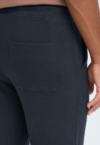 Pantalon de survêtement gris foncé en tissu texturé, avec une poche arrière unique et une coupe ajustée. Taille lisse avec des détails de couture subtils.