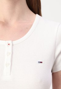 Valge ribaga lühikeste varrukatega pluus, millel on ümardatud kaelus, kolme nupuga klapp ja väike sinine, punane ning valge logo rinnal.