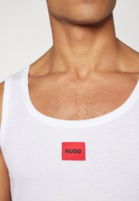 Biała bawełniana koszulka bez rękawów z czerwonym prostokątnym naszywką z napisem "HUGO" w kolorze czarnym. Żebrowana faktura, okrągły dekolt.