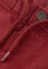 Tissu en coton bordeaux avec cordon tressé et détail de bouton en bronze. Les coutures visibles et la texture créent de la profondeur et de l'intérêt visuel.