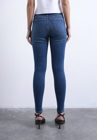 Γυναίκα που στέκεται με την πλάτη, φορώντας στενά μπλε τζιν, άσπρο τοπ και μαύρα ψηλοτάκουνα σανδάλια σε γκρίζο πάτωμα.