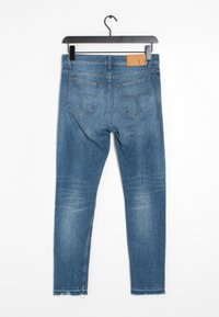Jeans de mezclilla azul expuestos colgando, con una cintura baja, diseño de cinco bolsillos y detalles de dobladillo sin terminar. Parche de logo de cuero en la parte posterior.