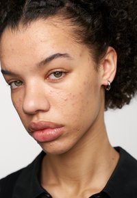 Portrait d'une personne à la peau brun clair, avec des cheveux courts et bouclés, et de l'acné. Elle a des lèvres pleines et porte une petite boucle d'oreille en argent, habillée en noir.