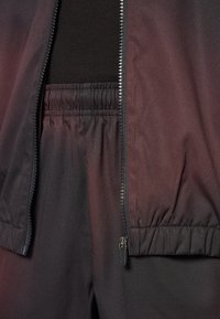 Melnā jaka ar rāvējslēdzēju priekšā un elastīgu jostasvietu. Tekstūrēta auduma ar nedaudz spīdumu, apvienota ar atbilstošām melnām elastīgām biksēm.