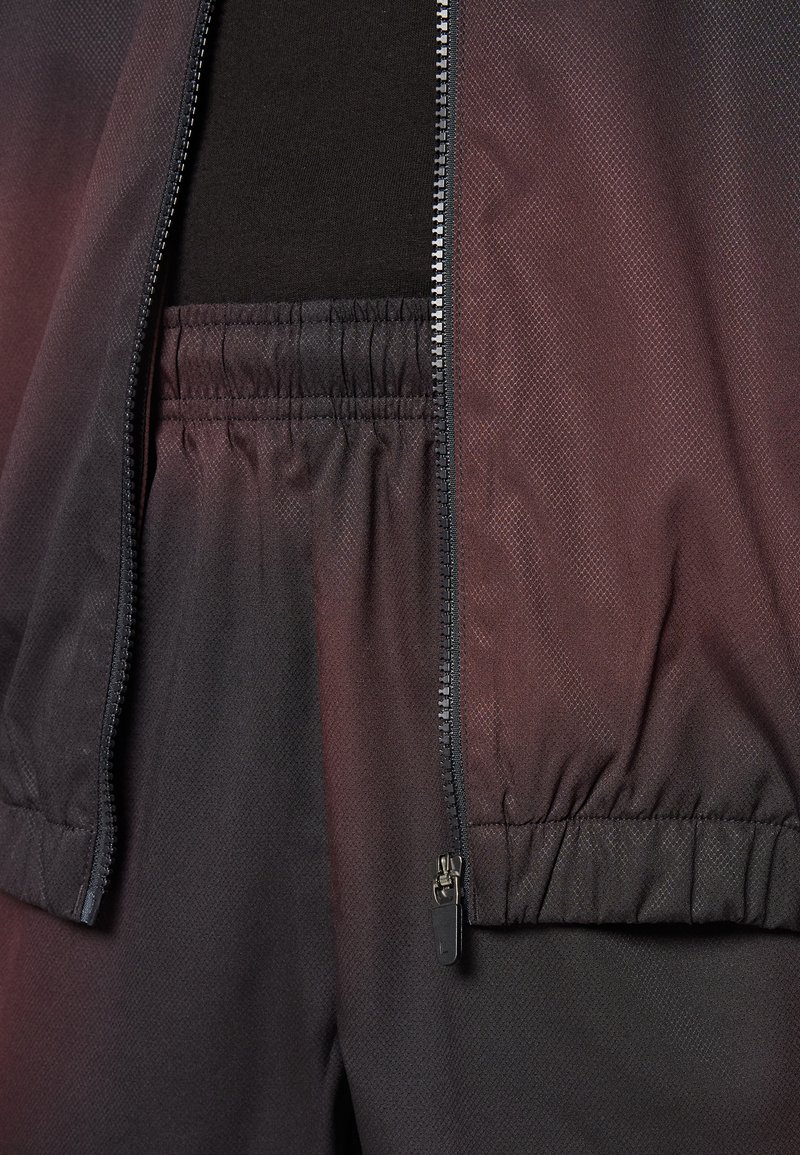 Schwarze Jacke mit Reißverschlussfront und elastischem Bund. Texturierter Stoff mit dezentem Glanz, kombiniert mit passenden schwarzen elastischen Hosen.