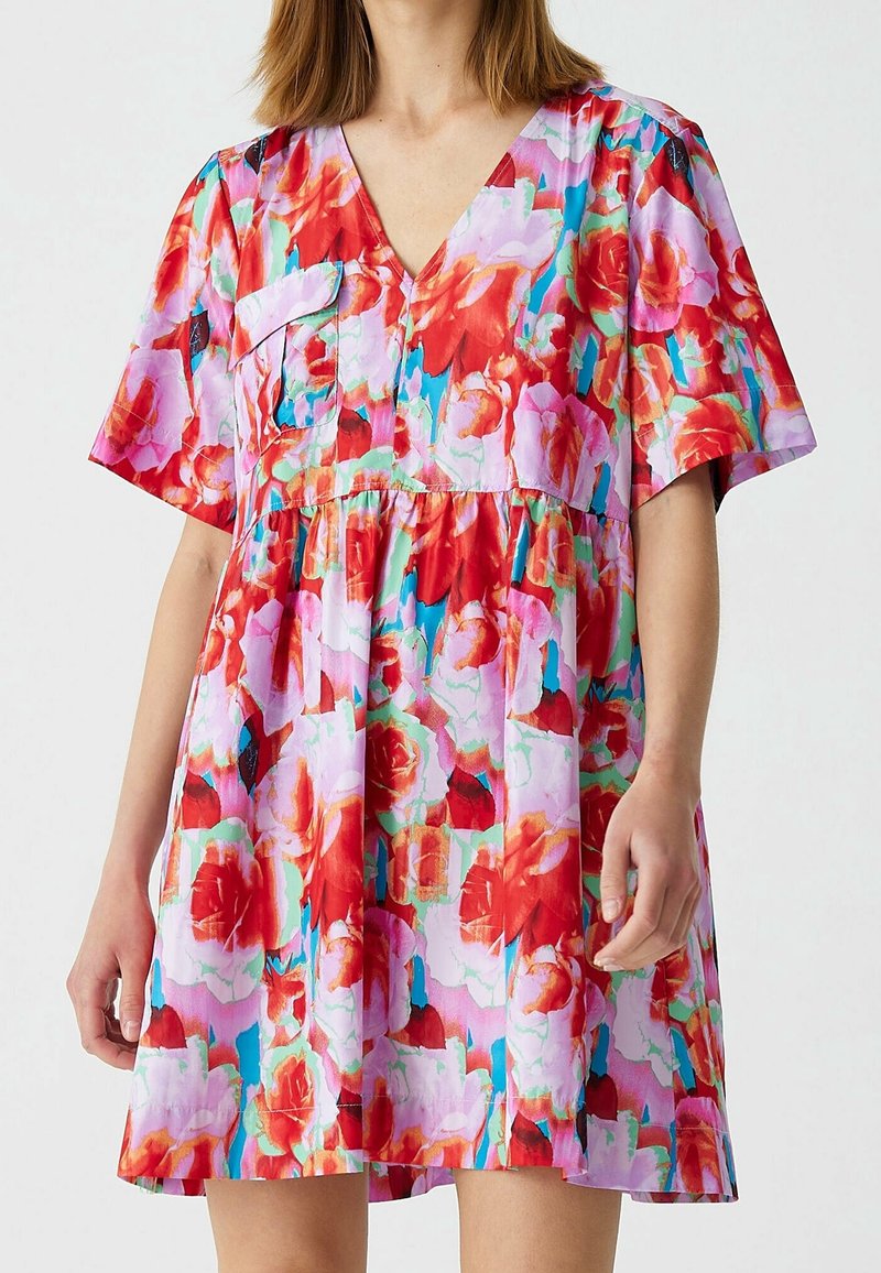 Femme portant une robe à manches courtes et col en V avec un imprimé floral vibrant rouge, rose et turquoise et une poche poitrine devant.