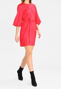 Horkorůžové krátké šaty s kulatým výstřihem a nabíranými rukávy, se zavázáním v pase. Model má na sobě černé kotníkové boty.