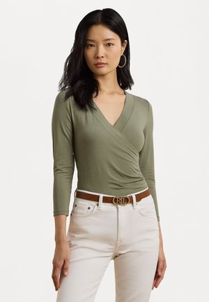 Haut enveloppant vert olive à manches longues et décolleté en V profond. Associé à un jean blanc et une ceinture marron avec un emblème doré.