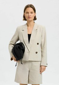 Lys beige dobbeltradet blazer og matchende shorts, lavet af en blanding af linned. Sort vævet taske med snøredetalje i hånden.