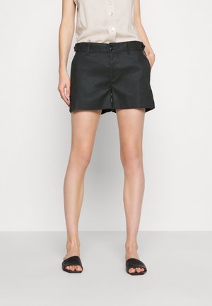 Frau trägt schwarze, maßgeschneiderte Shorts, beige ärmellose Bluse mit Knöpfen und schwarze offene Slip-On-Sandalen, steht vor einfachem Hintergrund.