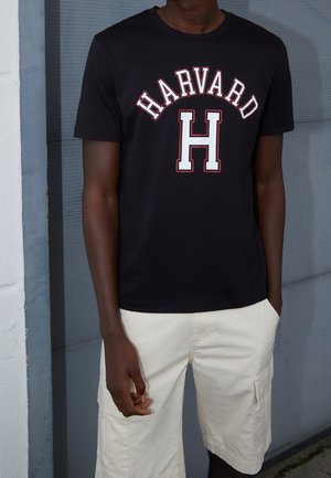 Person iført en sort t-shirt med teksten "HARVARD" og et stort "H" i hvidt og rødt, kombineret med lyse cargobermudaer.