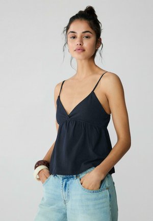 Fiatal, sötét hajú nő kontyba kötve, fekete pántos felsőt, világoskék farmert és vastag karkötőket visel, és zsebre tett kézzel pózol.