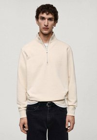 Cremfarbener Pullover mit Stehkragen aus weichem Stoff, mit einem halben Reißverschluss und gerippten Bündchen, kombiniert mit dunklen Jeans.