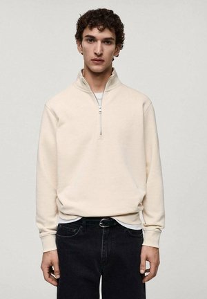 Cream-colored mock neck pullover made from soft fabric, featuring a half-zip closure and ribbed cuffs, paired with dark jeans.