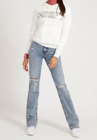 Felpa bianca con un logo argento sul petto, abbinata a jeans strappati azzurri chiari e sneakers bianche. Stile urbano casual.