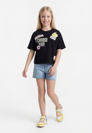 Νεαρή κοπέλα χαμογελά, φοράει μαύρο T-shirt Pokémon με το κείμενο «ΕΠΙΛΕΓΩ ΕΣΕΝΑ!», τζιν σορτς και πολύχρωμα αθλητικά παπούτσια, προχωράει μπροστά.