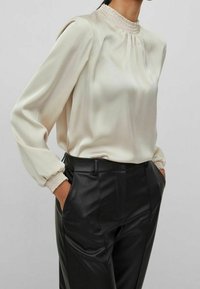 Blouse en satin crème avec col montant smocké et manches longues bouffantes, associée à un pantalon en cuir noir taillé sur mesure. Texture lisse, silhouette ajustée.