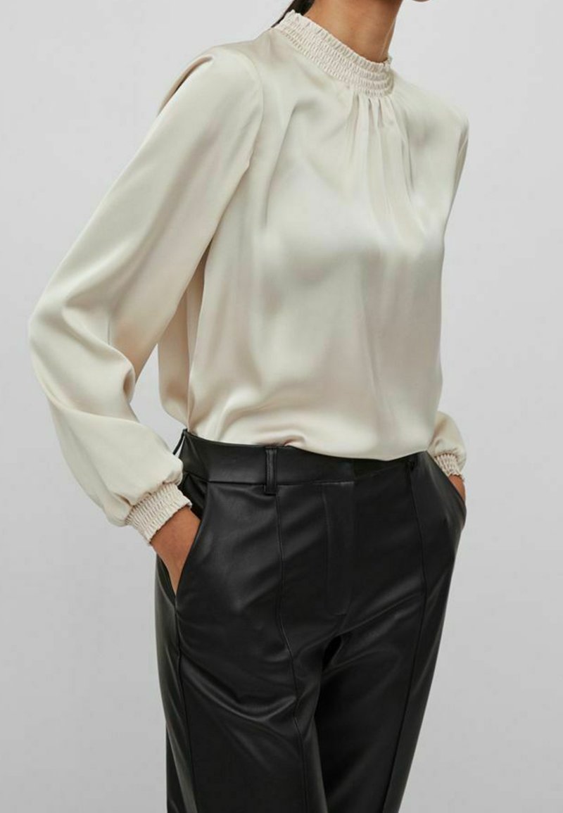 Blouse en satin crème avec col montant smocké et manches longues bouffantes, associée à un pantalon en cuir noir taillé sur mesure. Texture lisse, silhouette ajustée.