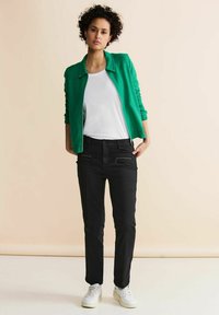 Grön zip-up jacka över en vit t-shirt, kombinerad med svarta byxor med dragkedjefickor och vita sneakers. Släta texturer genomgående.