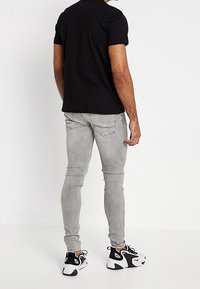 Hombre vestido con jeans gris ajustados, camiseta negra y zapatillas blancas y negras, de pie contra un fondo blanco liso mirando hacia atrás.