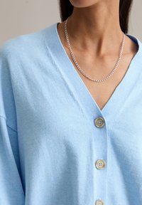Jasnoniebieski kardigan z dekoltem w serek, z czterema drewnianymi guzikami z przodu, miękka tekstura i luźny fason. Widoczny srebrny naszyjnik z łańcuszkiem.