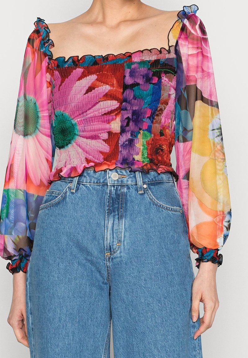 Mehrfarbige transparente Bluse mit floralem Muster, gerafftem Oberteil und Volantärmeln; kombiniert mit hochtaillierten blauen Jeans.
