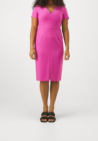 Femme portant une robe rose ajustée, mi-longue jusqu'aux genoux, à manches courtes, col en V, couture devant, et des sandales noires à bouts ouverts avec des brides volantées.