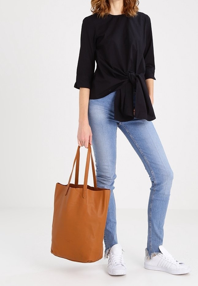 Personne portant un haut noir, un jean bleu clair, des baskets blanches, tenant un grand sac cabas en cuir marron par les poignées.
