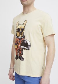Kortærmet, lys gul t-shirt med en tegneseriehund i rumdragtn med solbriller. Materialet ser blødt ud. Designet inkluderer klare farver.