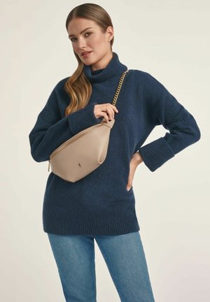 Granatowy sweter typu golf z prążkowanym dołem, w połączeniu z beżową torebką na ramię z zamkiem błyskawicznym i łańcuszkowym paskiem. Niebieskie dżinsy.