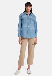 Camicia di denim azzurro chiaro con due tasche sul petto, chiusura con bottoni e maniche lunghe, abbinata a pantaloni cargo beige e sneaker bianche.