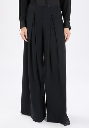 Donna che indossa pantaloni neri a gamba larga e vita alta con pieghe frontali, abbinati a una maglia nera a maniche lunghe infilata dentro e scarpe nere.