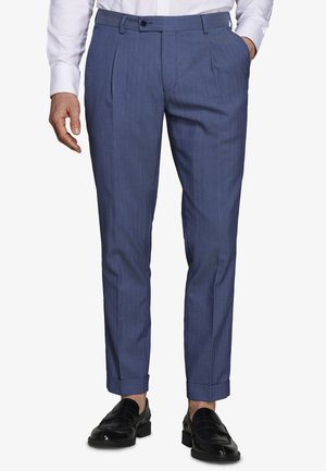 Homme portant un pantalon de costume bleu sur mesure, une chemise blanche et des mocassins en cuir noir, debout avec une main dans la poche, sur un fond blanc.
