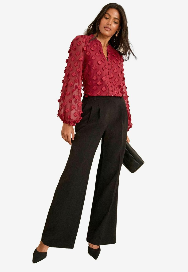 Blusa rossa trasparente con motivi floreali a texture e maniche lunghe a sbuffo, abbinata a pantaloni neri a gamba larga e scarpe nere a punta.