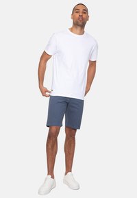 Hvid bomulds T-shirt parret med marineblå shorts. Model iført hvide sneakers, står med den ene hånd på shortsene, afslappet pasform.