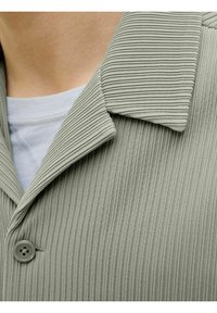 Chaqueta de color verde claro, con canalé y un acabado texturizado, que presenta un cuello con muesca y un cierre de un solo botón. Camiseta de cuello redondo azul visible por debajo.