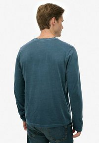 Lange mouw teal katoenen T-shirt met een ronde halslijn, een zachte textuur en een licht beschadigde zoom, gedragen met donkere jeans.