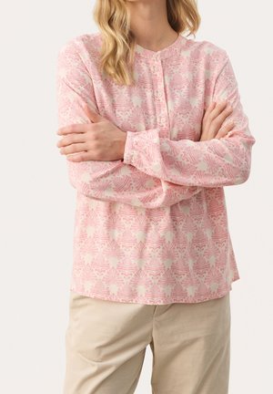 Kobieta ubrana w różową, wzorzystą bluzkę z długim rękawem i guzikami, beżowe spodnie, z założonymi na piersi skrzyżowanymi ramionami, na jednolitym tle.