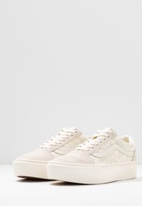 Niskie beżowe trampki z zamszowymi noskami, tonowanymi sznurówkami oraz subtelnym wzorzystym materiałem górnym, na którym powtarza się tekst logo.