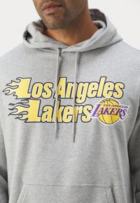 Felpa grigia con cappuccio che presenta il testo "Los Angeles Lakers" in giallo e nero insieme a una grafica di un pallone da basket. Il materiale sembra morbido e dotato di un cappuccio regolabile con cordino.