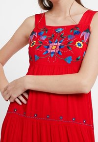 Robe rouge sans manches avec broderies florales en bleu, rose et jaune sur le corsage, dotée d'une jupe froncée et d'accents bleus le long de l'ourlet.