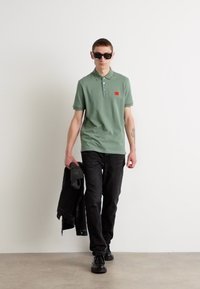 Grønn polo-skjorte med korte ermer, rød lappedetalj, svarte jeans, bærer en svart jakke og har på seg svarte solbriller. Glatt tekstur.