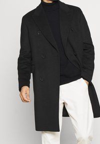 Abrigo de lana negro de doble botonadura con solapas muesca, dos bolsillos frontales y botones grandes. Llevado sobre un cuello de tortuga negro, combinado con pantalones blancos.