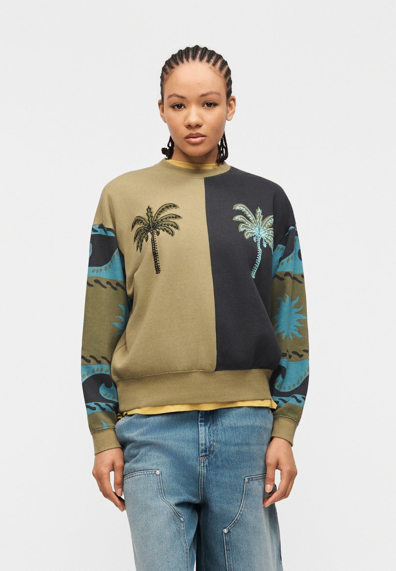 Νεαρό άτομο με πλεξούδες φορώντας φούτερ διχρωμίας με σχέδια φοίνικα και μανίκια με μπλε μοτίβα, σε συνδυασμό με ανοιχτόχρωμα μπλε τζιν.