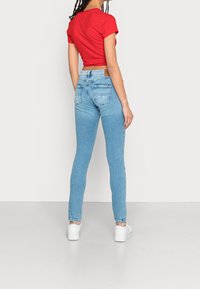 Mujer vestida con jeans ajustados de color azul claro, camiseta corta de manga corta roja, zapatillas blancas, de pie mirando hacia atrás contra un fondo claro.