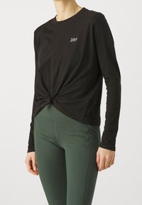 Svart långärmad tröja med knutdetalj framtill och broderad "DKNY"-logotyp, tillsammans med gröna leggings. Slät tygtextur.